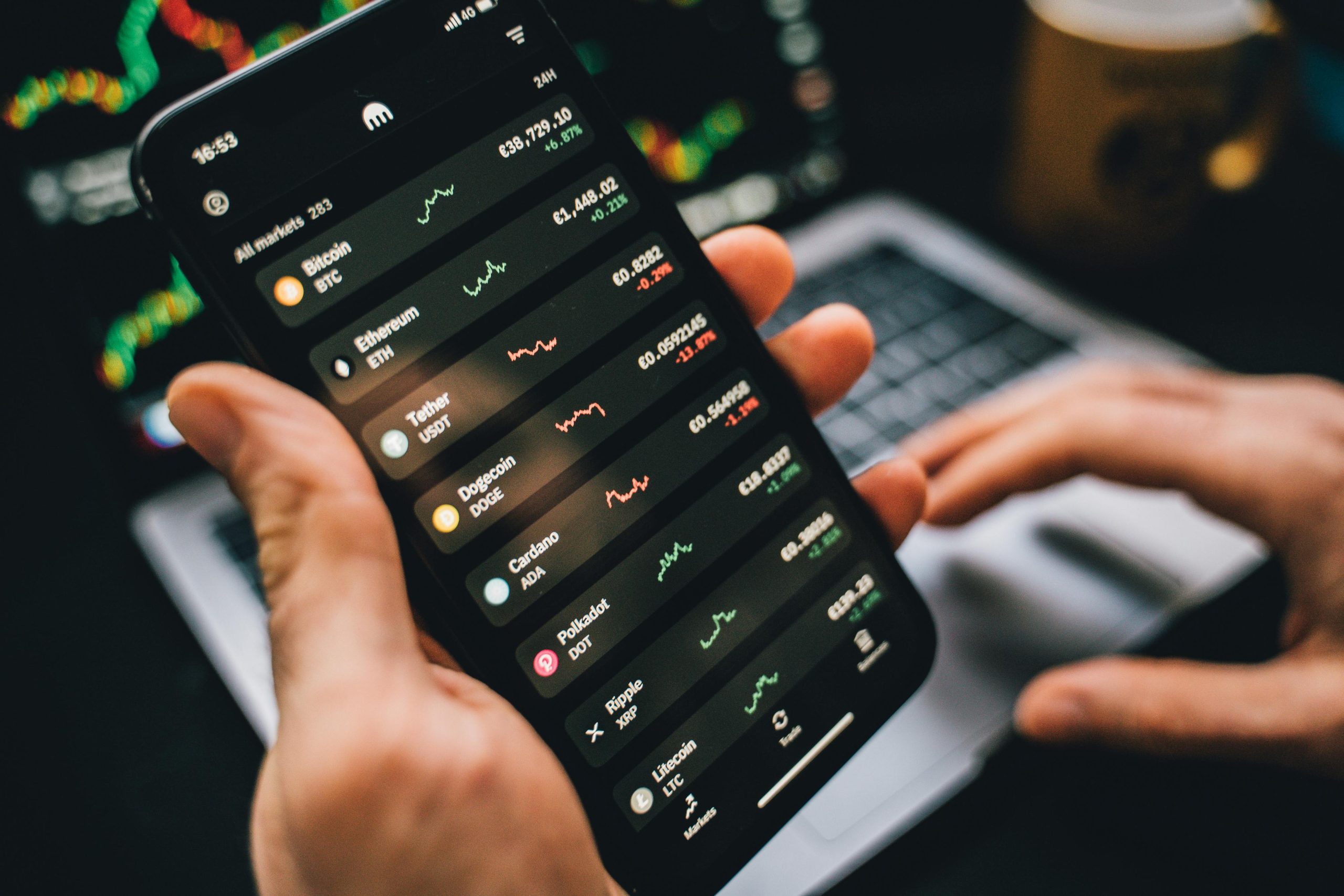 A close-up view of a hand holding a smartphone displaying a cryptocurrency market app. The screen shows various cryptocurrencies like Bitcoin, Ethereum, and Dogecoin, with price changes and market trends in green and red. A laptop and a coffee mug are visible in the background.