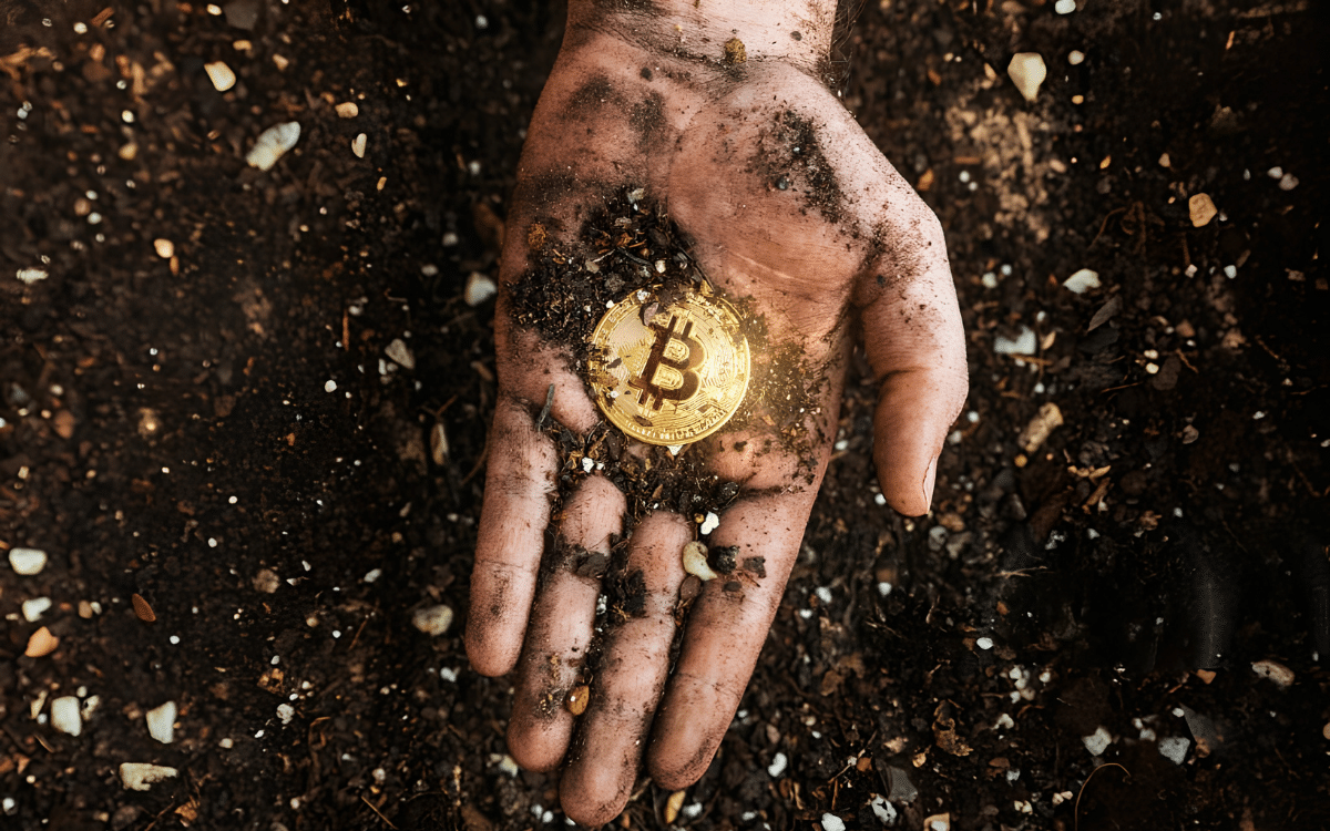 A dirty hand holding a gold Bitcoin coin in soil, illustrating Bitcoin mining.