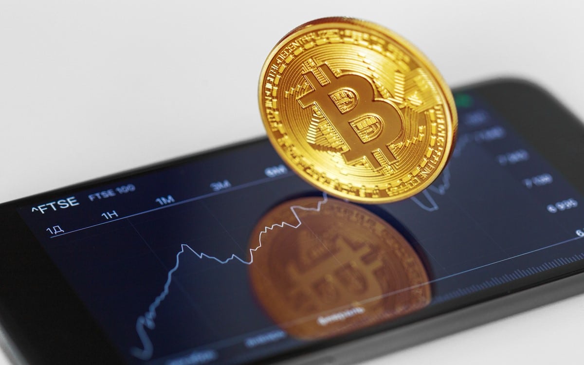 An image reveals a Bitcoin coin laying on top of a smartphone showing a crypto price chart on the phone.